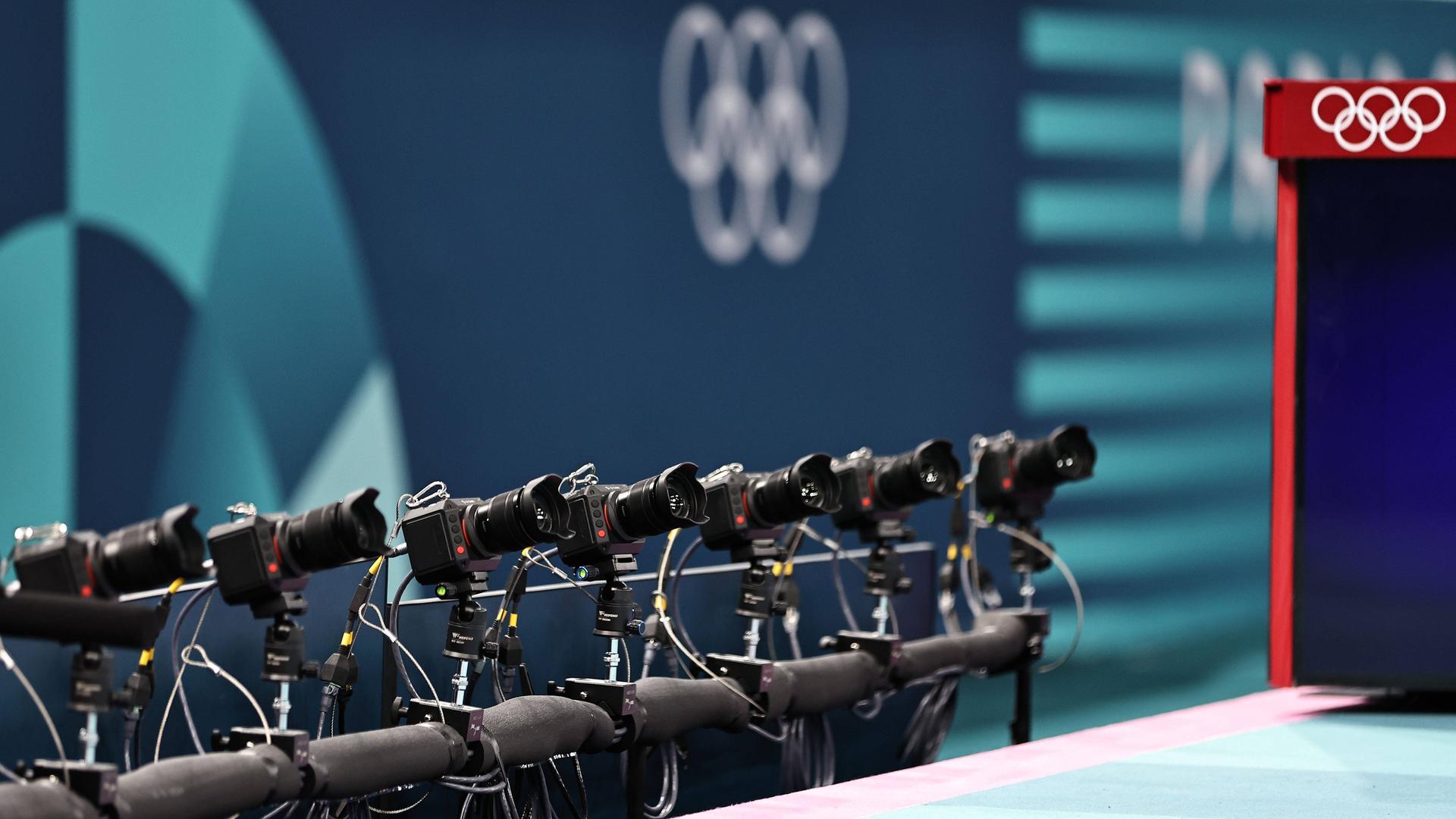 Zu sehen ist eine lange Reihe an Kameras, die auf eine Turn-Wettbewerbsfläche gerichtet sind, im Hintergrund sind die Olympischen Ringe.