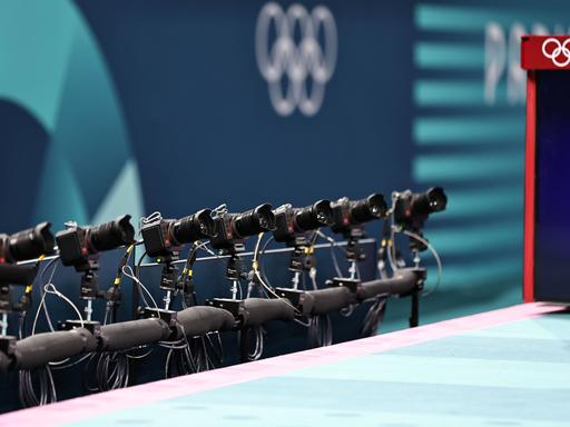 Zu sehen ist eine lange Reihe an Kameras, die auf eine Turn-Wettbewerbsfläche gerichtet sind, im Hintergrund sind die Olympischen Ringe.