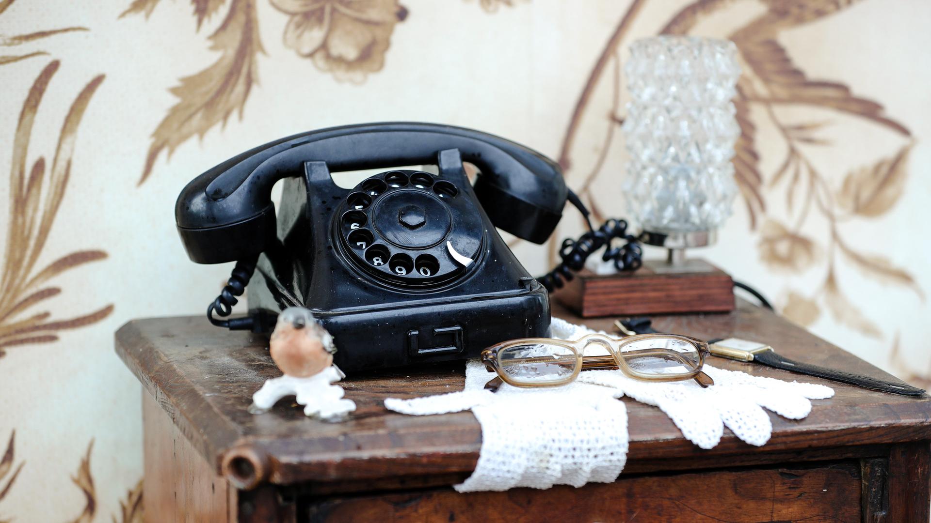 Ein schwarzes, altmodisches Telefon mit Wählscheibe auf einer Kommode mit Spitzendeckchen. 