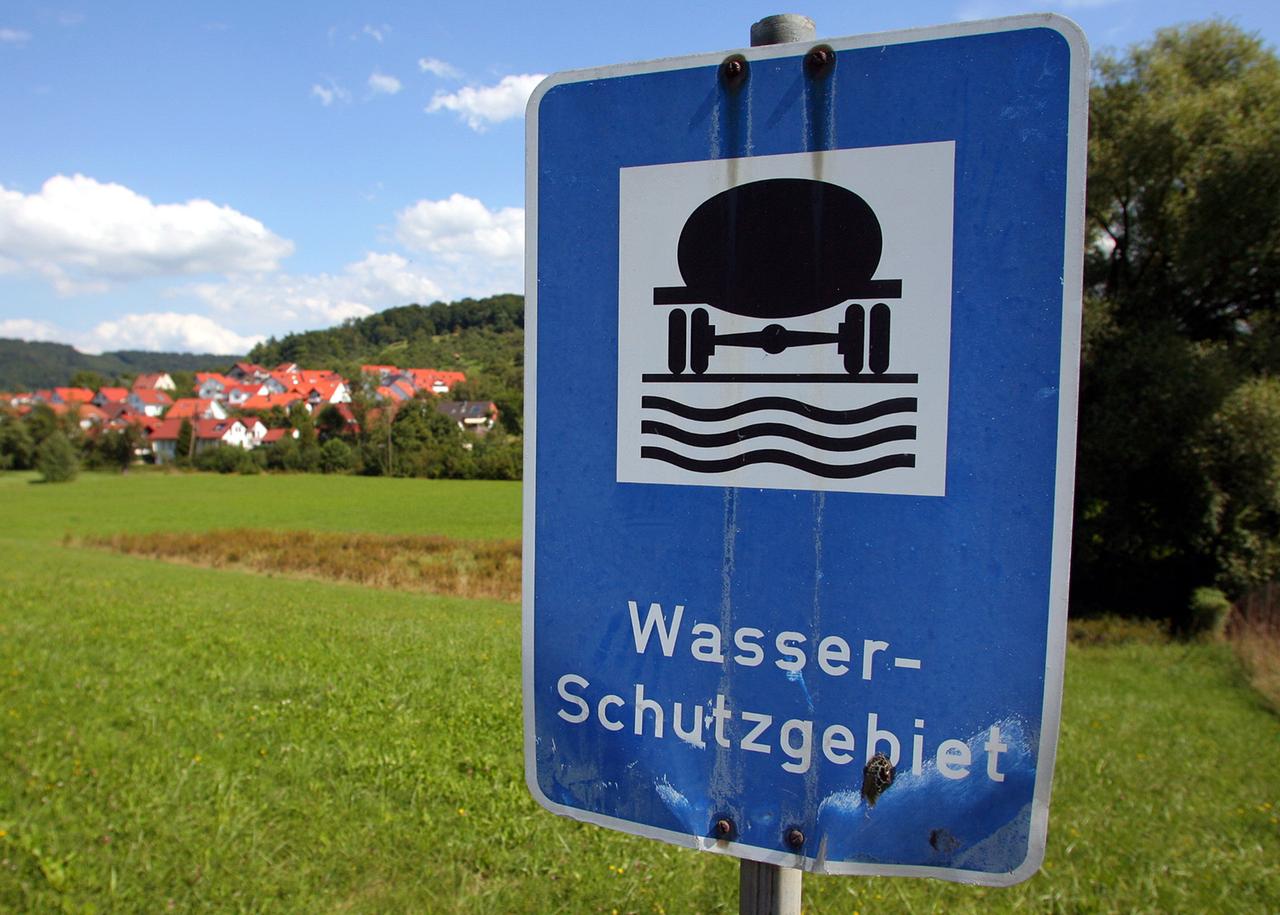 Ein blaues Schild mit der Aufschrift "Wasserschutzgebiet" steht vor einem Feld, im Hintergrund sind einige Häuser zu sehen.