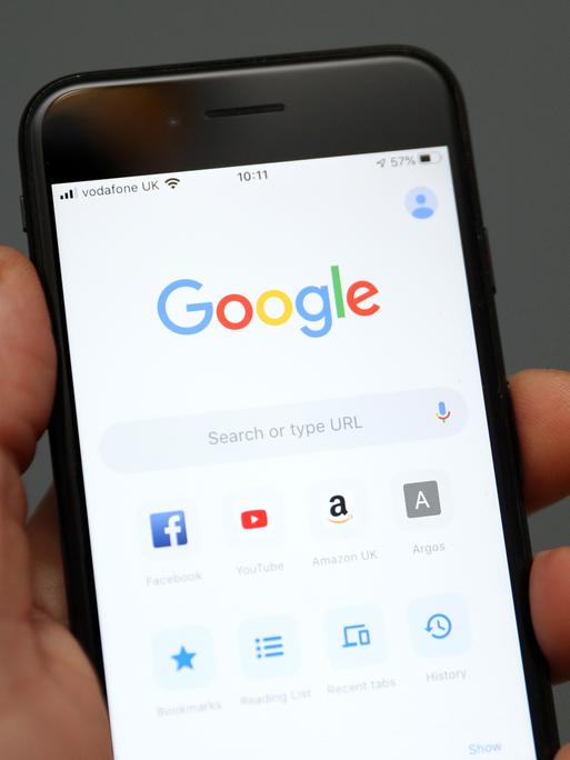 Eine Hand hält ein Mobiltelefon auf dessen Display die Oberfläche der Suchmaschine Google zu sehen ist. 