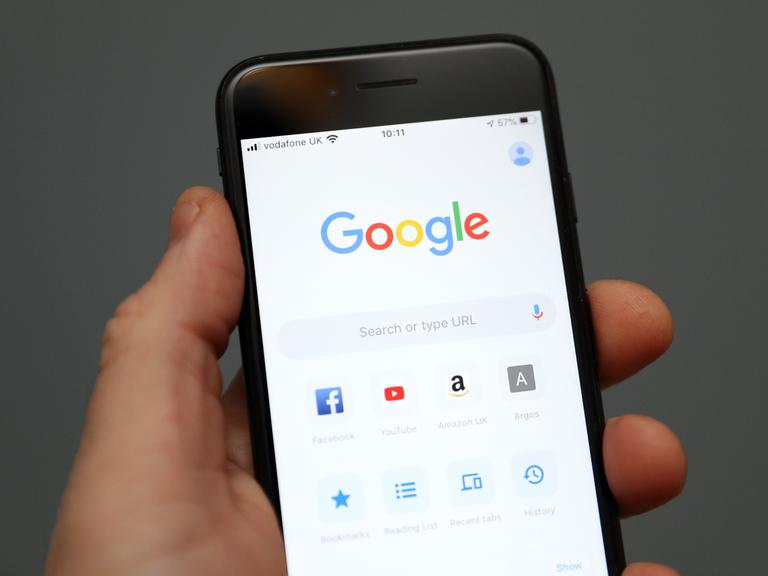Eine Hand hält ein Mobiltelefon auf dessen Display die Oberfläche der Suchmaschine Google zu sehen ist. 