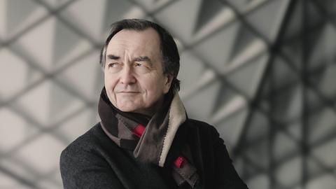 Ein Mann mit dunklen Haaren steht mit verschränkten Armen vor einer Glaswand mit Dreiecksmuster. Er schaut in die Ferne, lächelt verschmitzt. Er trägt einen dunklen Mantel und einen Wollschal. 