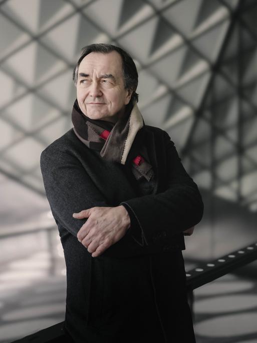 Ein Mann mit dunklen Haaren steht mit verschränkten Armen vor einer Glaswand mit Dreiecksmuster. Er schaut in die Ferne, lächelt verschmitzt. Er trägt einen dunklen Mantel und einen Wollschal. 