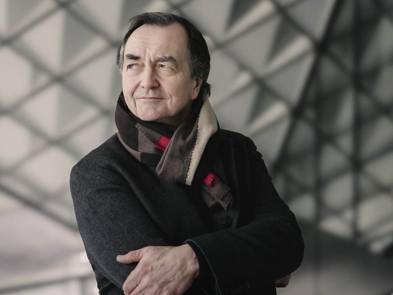 Ein Mann mit dunklen Haaren steht mit verschränkten Armen vor einer Glaswand mit Dreiecksmuster. Er schaut in die Ferne, lächelt verschmitzt. Er trägt einen dunklen Mantel und einen Wollschal. 