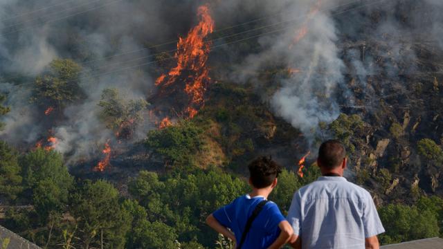 Zwei Personen blicken auf einen brennenden bewaldeten Berghang. Zwischen grünen Bäumen schlagen an mehreren Stellen hohe orangefarbene Flammen empor. Dichte graue Rauchschwaden ziehen über den Hang, mehrere Stromleitungen verlaufen durch das Bild.