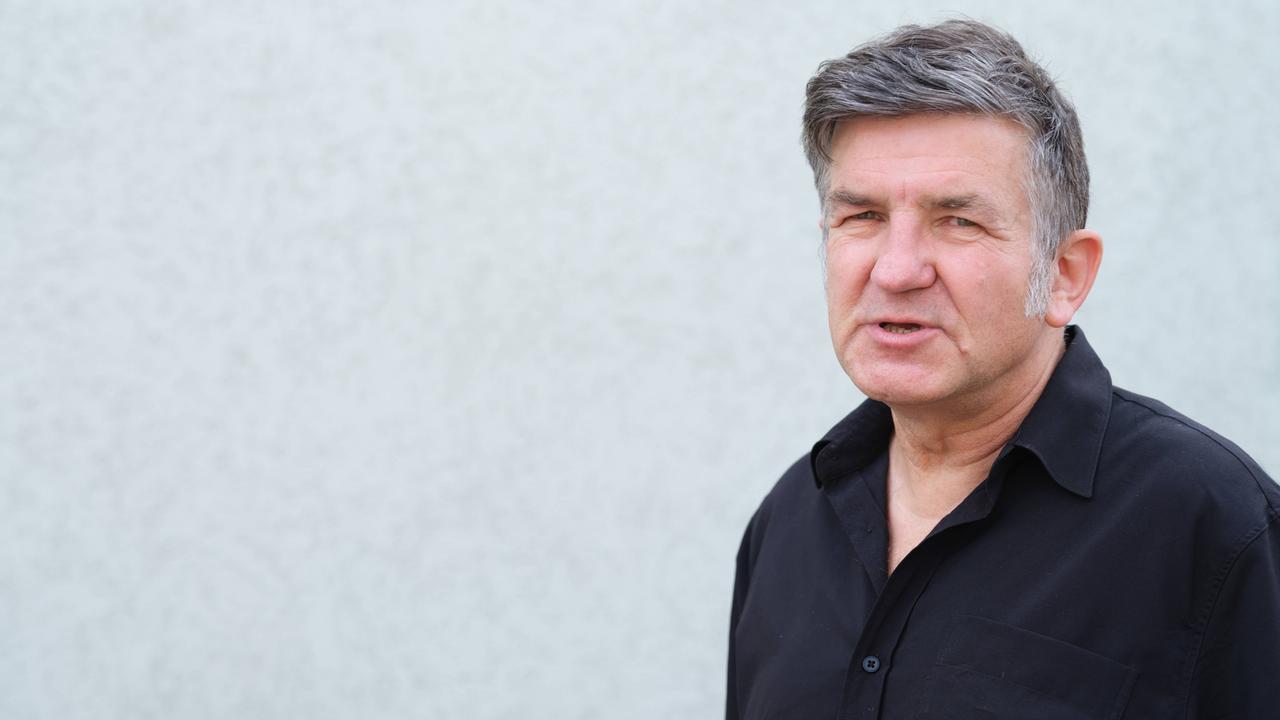 Ein Mann mit grauer Tolle und Koteletten steht vor einer Wand. Er trägt ein schwarzes Hemd.