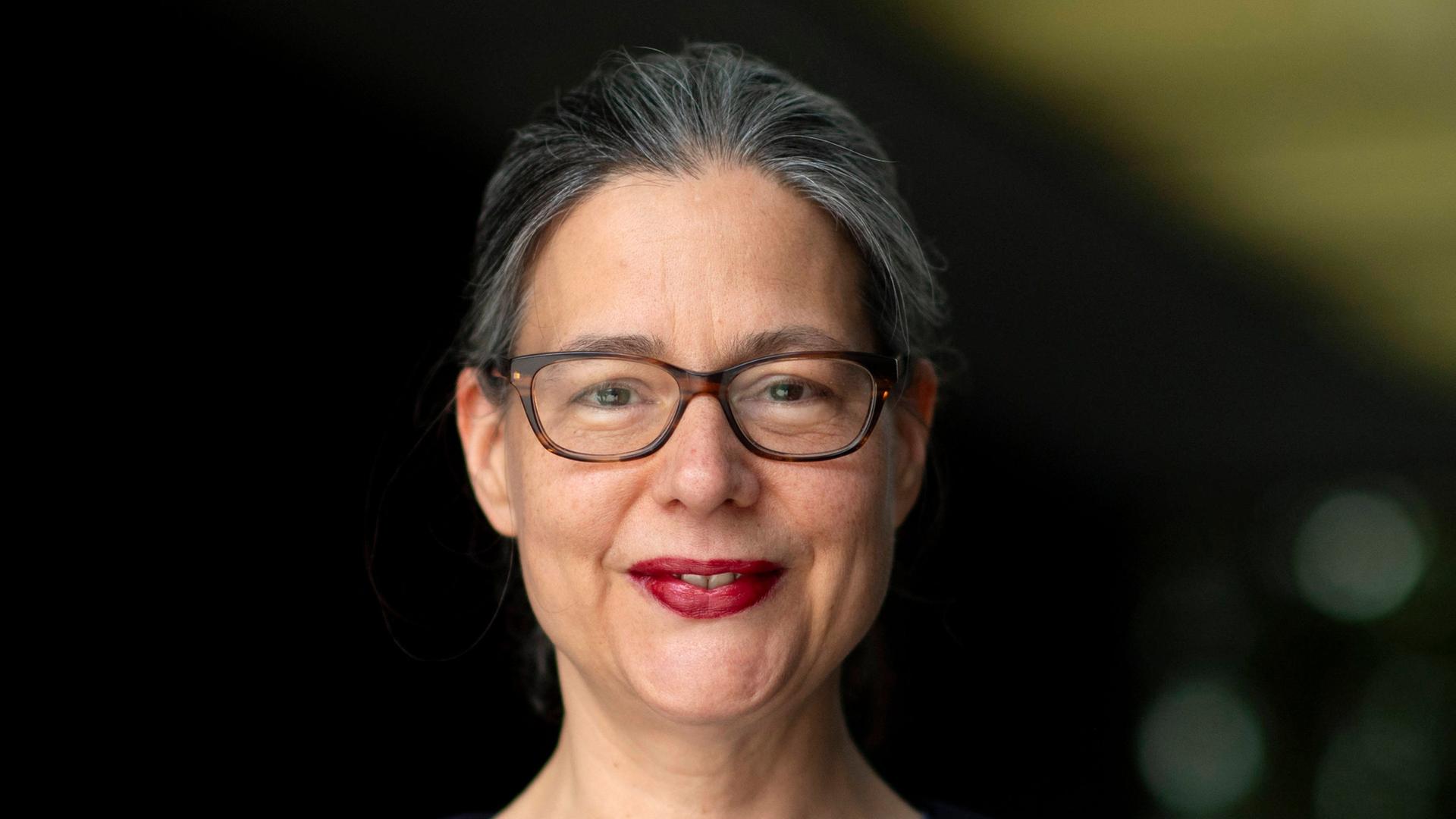 Porträt von Nina Scheer (SPD). Sie guckt vor dunklem unscharfem Hintergrund in die Kamera und lächelt. Sie trägt eine Brille und roten Lippenstift. Dazu eine blaue Jacke über einem grauen Oberteil.