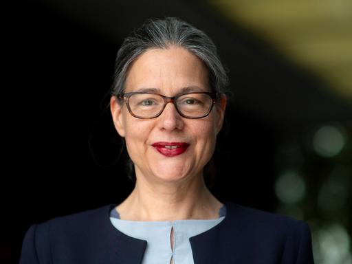 Porträt von Nina Scheer (SPD). Sie guckt vor dunklem unscharfem Hintergrund in die Kamera und lächelt. Sie trägt eine Brille und roten Lippenstift. Dazu eine blaue Jacke über einem grauen Oberteil.