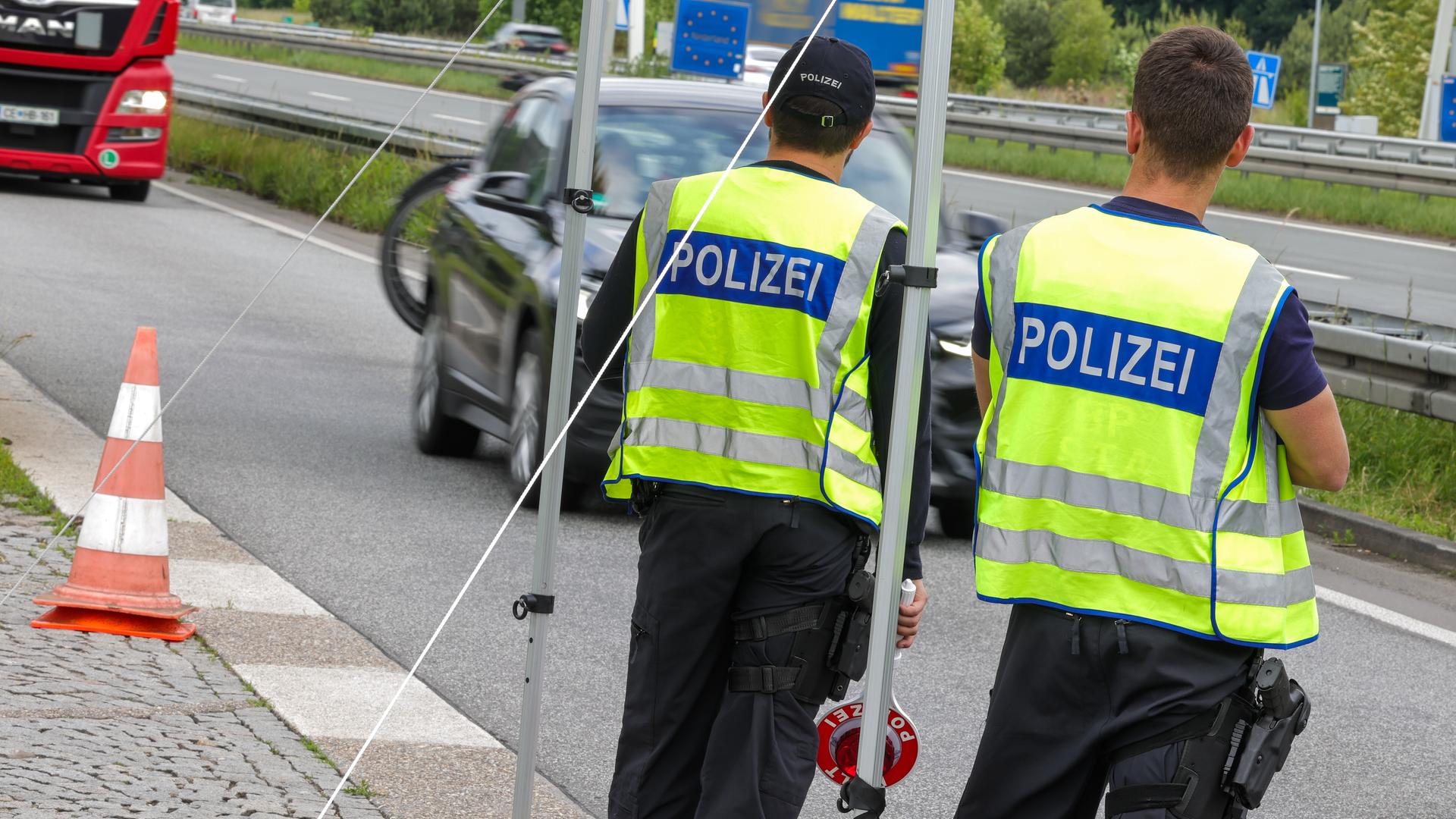 An der Grenze zu den Niederlanden kontrollieren zwei Bundespolizisten die nach Deutschland einfahrenden Fahrzeuge. Sie tragen gelbe Westen mit der Aufschrift Polizei und sind nur von hinten abgebildet.  