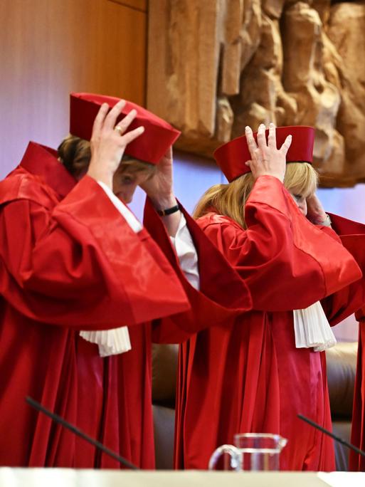 Richterinnen und Richter des Bundesverfassungsgerichts stehen vor einer Verhandlung in roten Roben hinter ihrem Tisch und setzen sich rote Mützen auf.