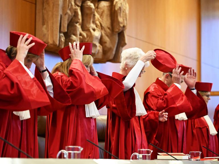 Richterinnen und Richter des Bundesverfassungsgerichts stehen vor einer Verhandlung in roten Roben hinter ihrem Tisch und setzen sich rote Mützen auf.