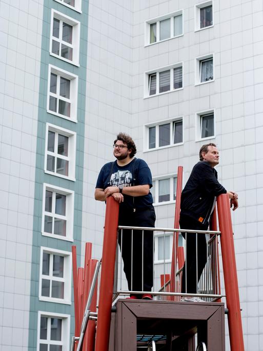 Devid Striesow und Axel Ranisch stehen auf einem Klettergerüst vor einer Plattenbaufassade und schauen in unterschiedliche Richtungen.