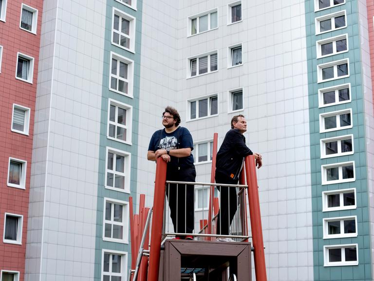 Devid Striesow und Axel Ranisch stehen auf einem Klettergerüst vor einer Plattenbaufassade und schauen in unterschiedliche Richtungen.