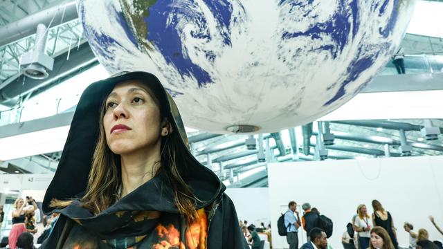 Eine kostümierte Demonstrantin steht mit düsterer Miene unter einer großen blauen Erdkugel in einer Messehalle, im Hintergrund sind weitere Teilnehmer der Weltklimakonferenz zu sehen.
