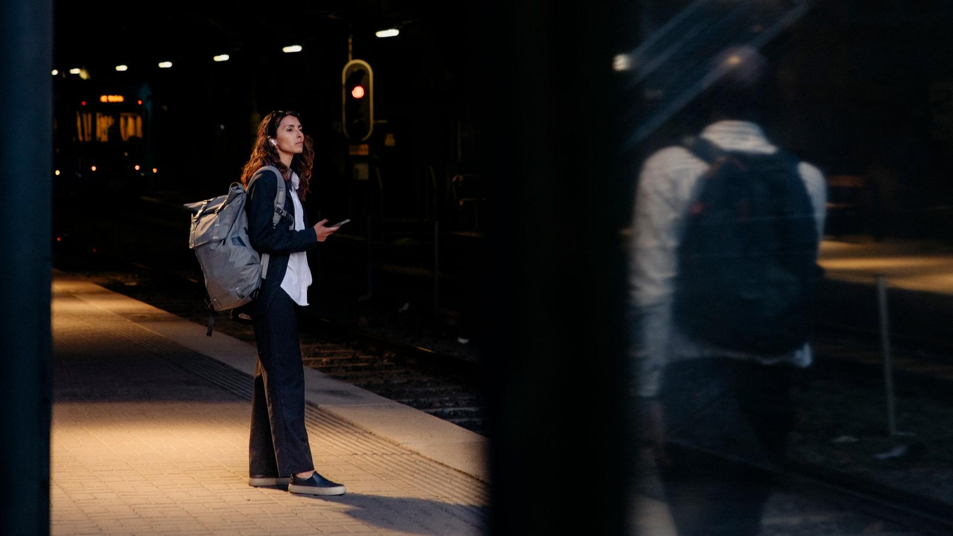 Eine Frau steht an einem Bahnhof. Unscharf im Bildvordergrund steht ein Mann, der sie beobachtet. Eine Frau steht an einem Bahnhof. Unscharf im Bildvordergrund steht ein Mann, der sie beobachtet.