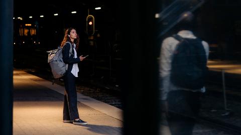 Eine Frau steht an einem Bahnhof. Unscharf im Bildvordergrund steht ein Mann, der sie beobachtet.