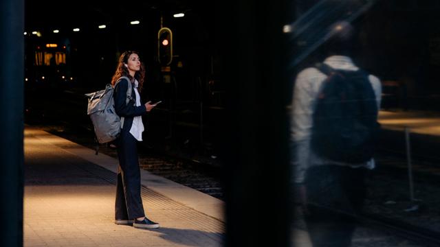Eine Frau steht an einem Bahnhof. Unscharf im Bildvordergrund steht ein Mann, der sie beobachtet.