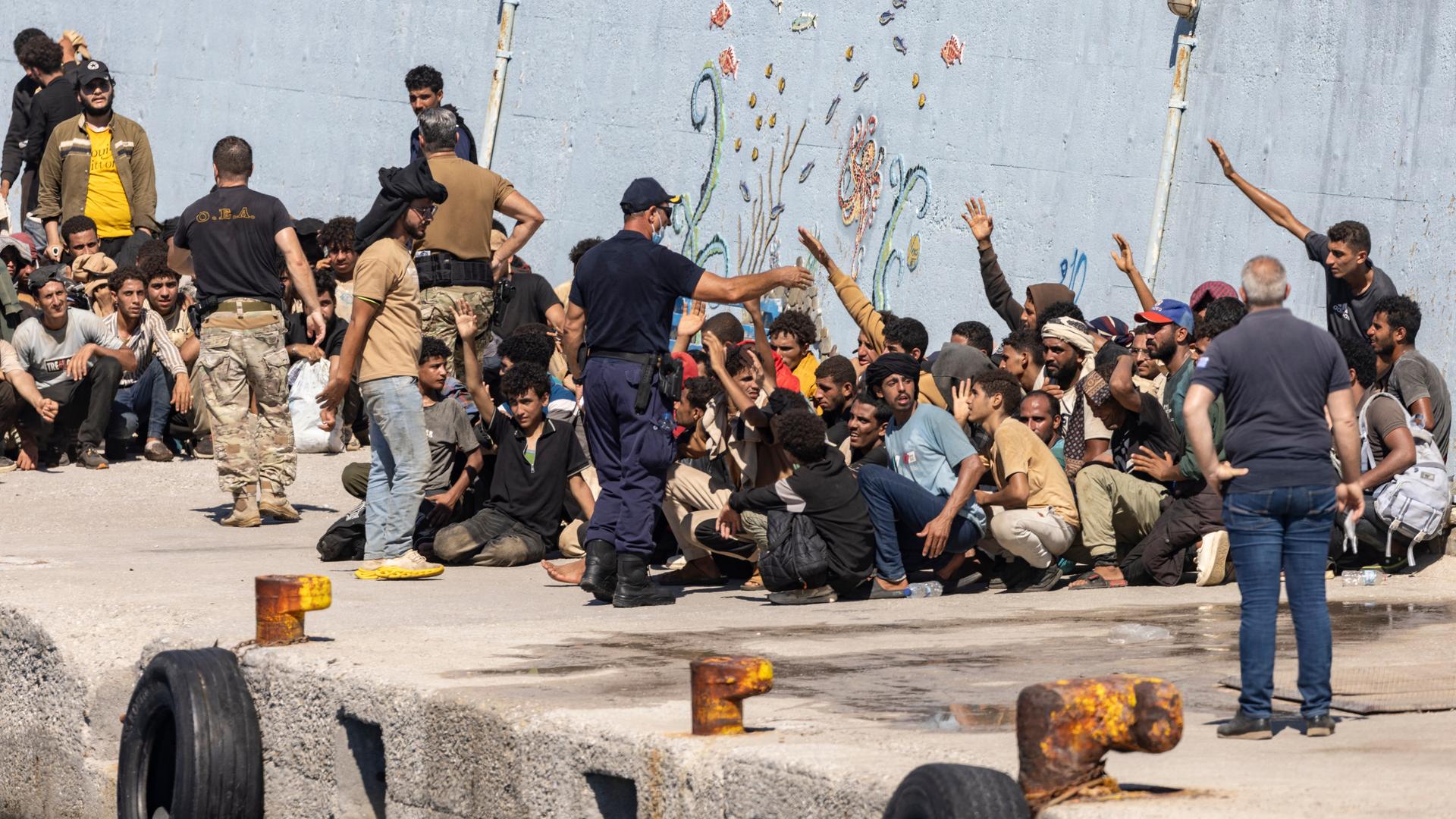 Migranten sitzen an einem Kai, einige von ihnen heben einen Arm. Um sie herum stehen Männer, bei einem von ihnen handelt es sich um einen Sicherheitsbeamten.
