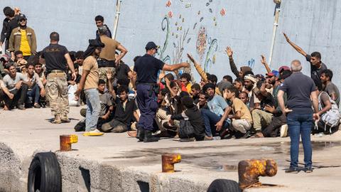 Migranten sitzen an einem Kai, einige von ihnen heben einen Arm. Um sie herum stehen Männer, bei einem von ihnen handelt es sich um einen Sicherheitsbeamten.