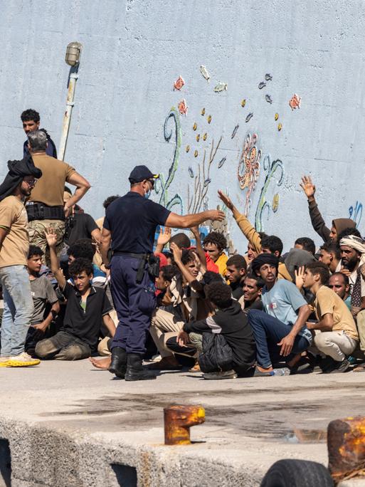 Migranten sitzen an einem Kai, einige von ihnen heben einen Arm. Um sie herum stehen Männer, bei einem von ihnen handelt es sich um einen Sicherheitsbeamten.
