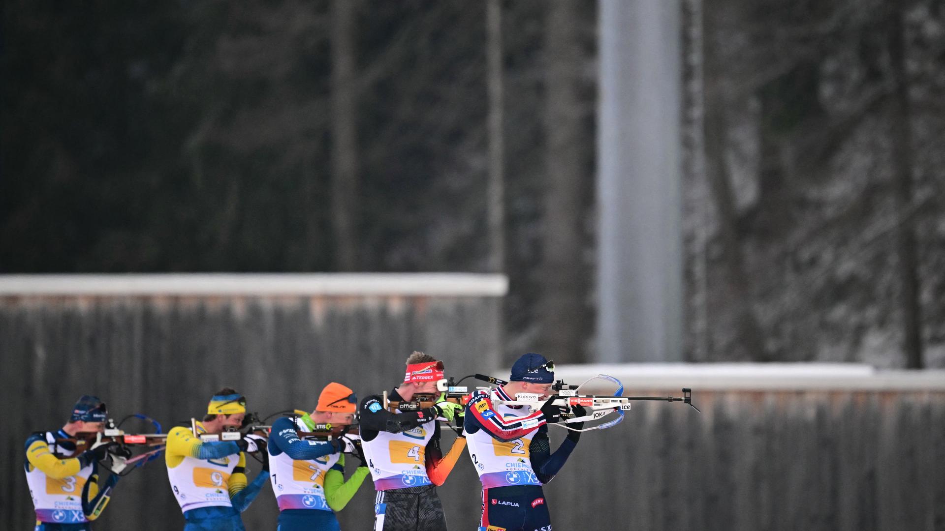 Blick aus der Ferne: Mehrere Biathleten stehen nebeneinander am Schießstand und haben ihre Gewehr im Anschlag. Dahinter eine Holzwand und Wald.