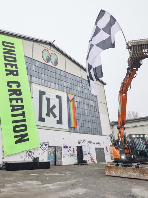 Ein Bagger schwenkt eine Schwarz-weiß karierte Zielflagge während einer künstlerischen Intervention von Daniel Dominguez Teruel auf der Piazza am Kampnagel anlässlich des Spatenstiches zur Sanierung und Erweiterung von Kampnagel. 