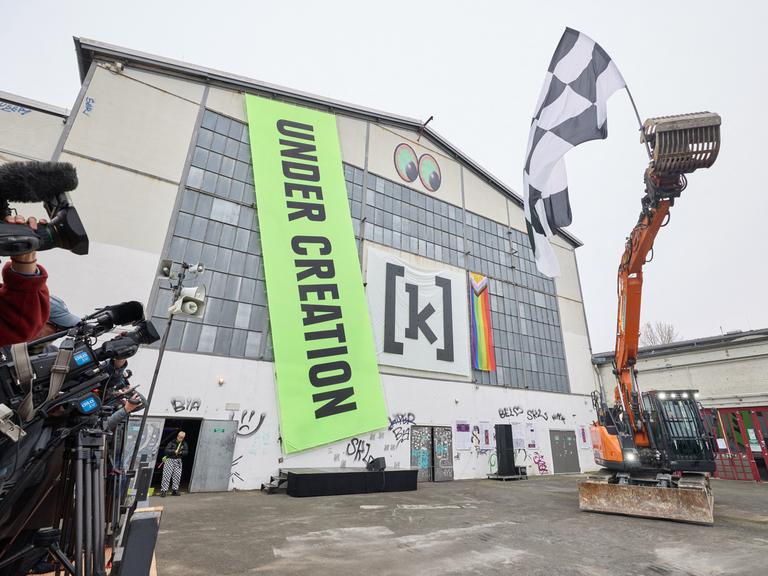 Ein Bagger schwenkt eine Schwarz-weiß karierte Zielflagge während einer künstlerischen Intervention von Daniel Dominguez Teruel auf der Piazza am Kampnagel anlässlich des Spatenstiches zur Sanierung und Erweiterung von Kampnagel. 