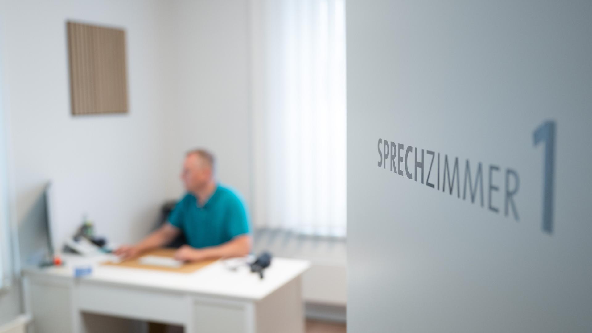 Ein Hausarzt sitzt in einem Sprechzimmer seiner Hausarztpraxis an einem Schreibtisch und arbeitet am Computer. 