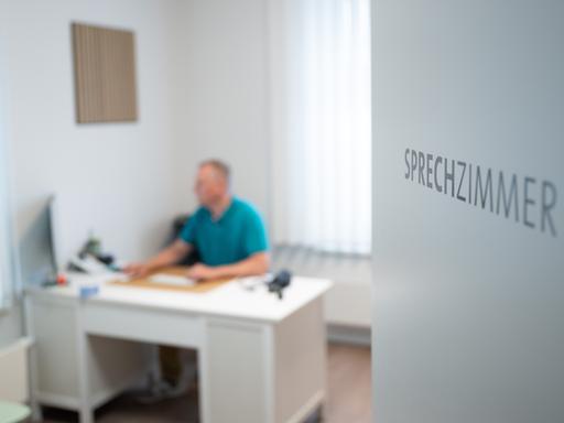 Ein Hausarzt sitzt in einem Sprechzimmer seiner Hausarztpraxis an einem Schreibtisch und arbeitet am Computer. 