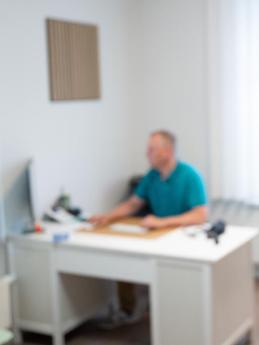 Ein Hausarzt sitzt in einem Sprechzimmer seiner Hausarztpraxis an einem Schreibtisch und arbeitet am Computer. 