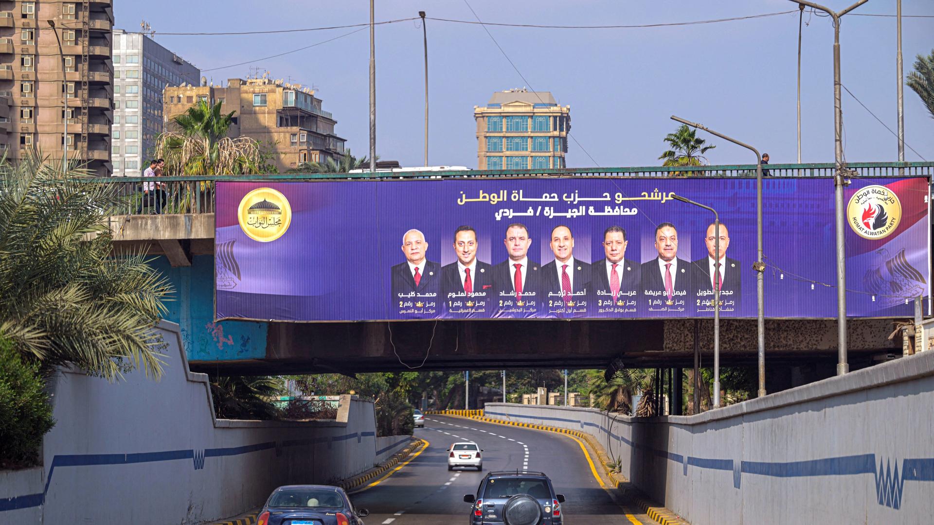 Das Bild zeigt eine Straßenszene in Ägypten. Autos fahren auf eine Brücke zu, an der ein Banner mit den Kandidaten zur Unterhauswahl hängt. Das Bild zeigt eine Straßenszene in Ägypten. Autos fahren auf eine Brücke zu, an der ein Banner mit den Kandidaten zur Unterhauswahl hängt.