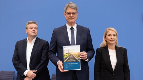 Die drei stehen nebeneinander vor der blauen Wand der Bundespressekonferenz. Schnieder steht in der Mitte und hält das Konzeptpapier in den Händen. Palla steht rechts von ihm. 