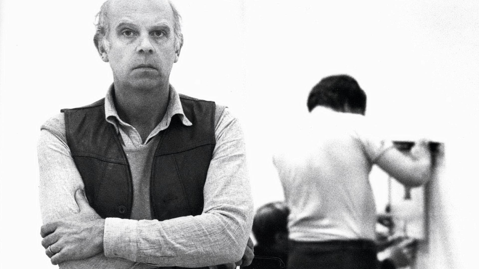 Schwarzweißaufnahme des Künstlers Claes Oldenburg, der mit ernstem Blick und verschränkten Armen in die Kamera blickt.