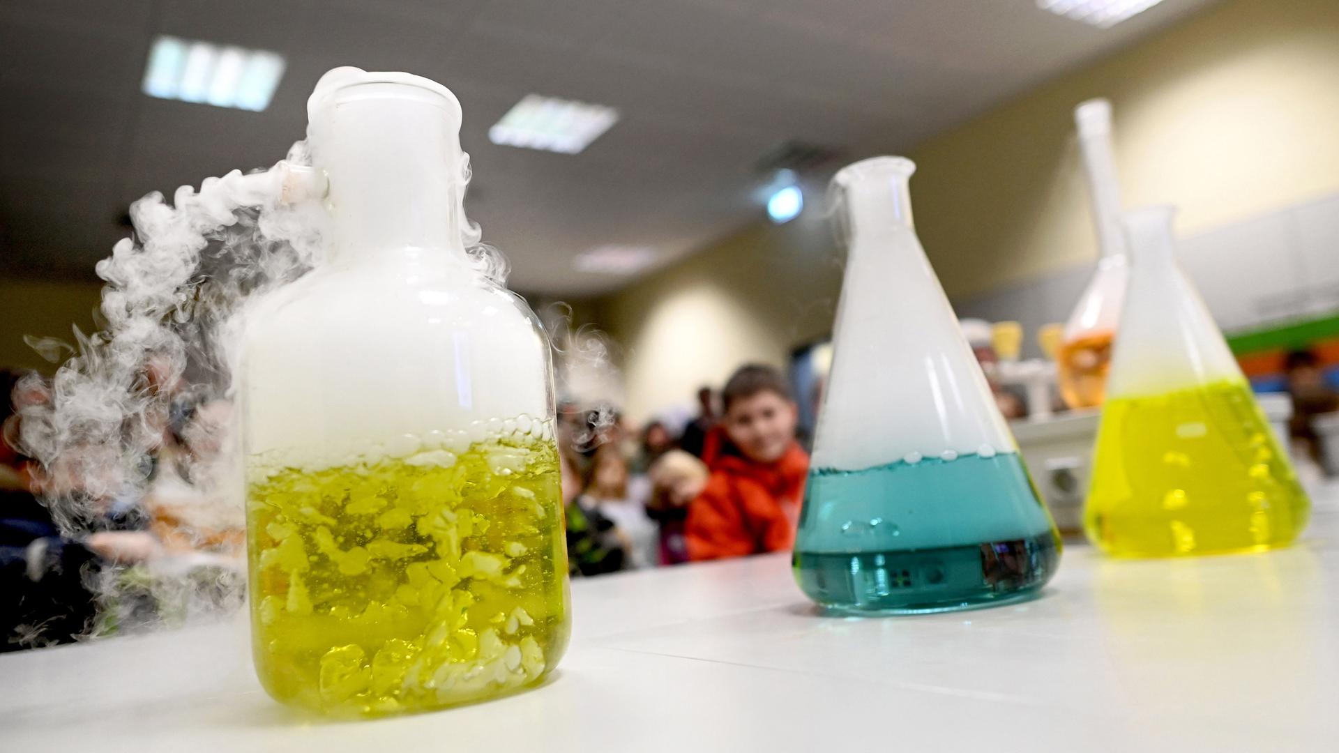 Mehrere Erlenmeyerkolben mit gefärbtem Wasser und Trockeneiswürfeln stehen auf einem Tisch im Chemieraum einer Schule. 