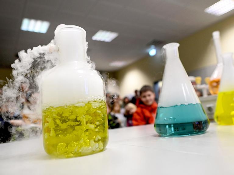 Mehrere Erlenmeyerkolben mit gefärbtem Wasser und Trockeneiswürfeln stehen auf einem Tisch im Chemieraum einer Schule. 