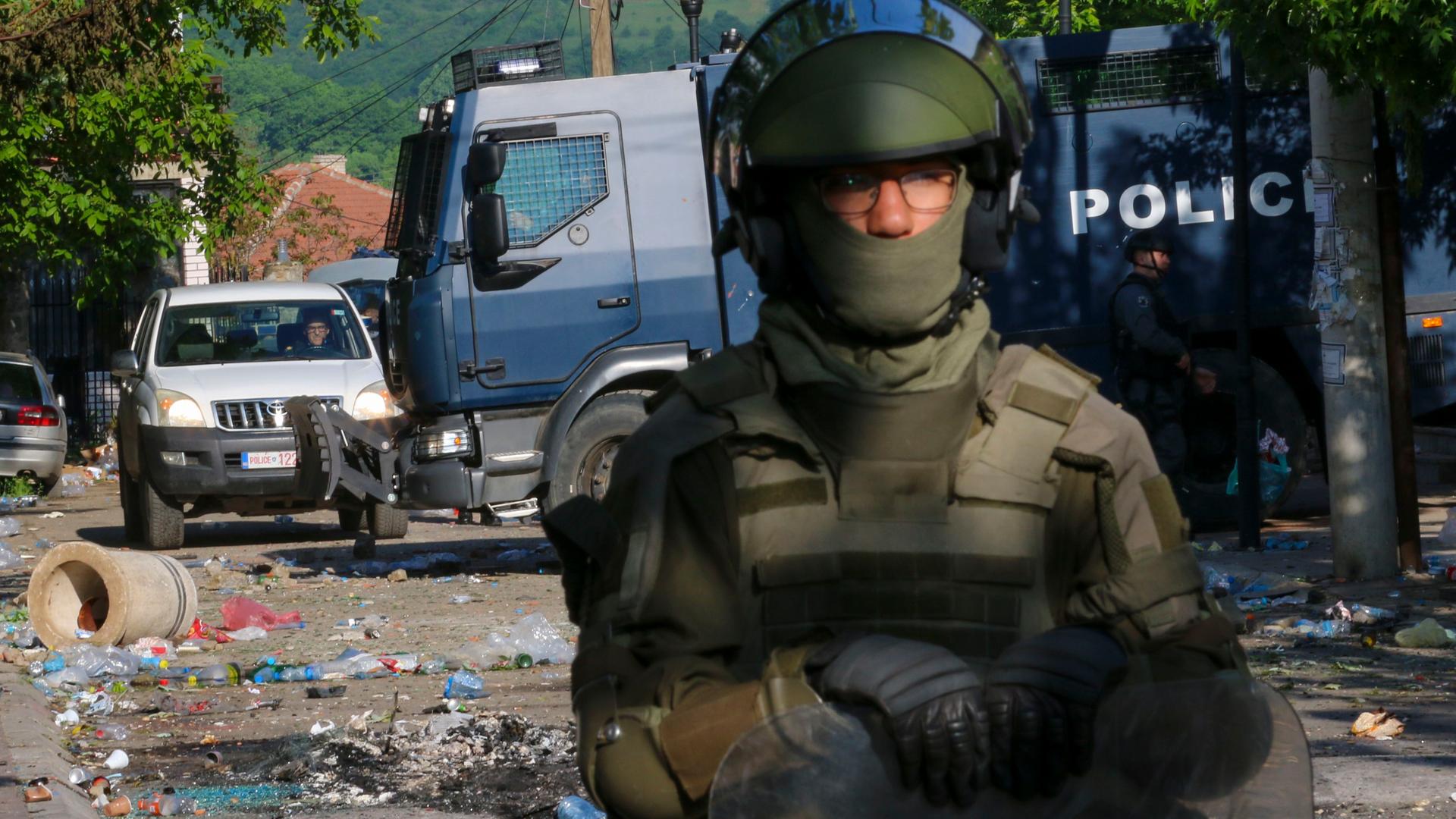 Ein KFOR-Soldat steht im Vordergrund. Hinter ihm zerstörte Autos und Trümmerteile auf der Straße im Kosovo.