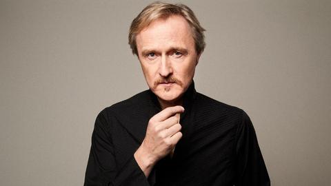 Albrecht Mayer steht im dunklen Rolli und erhobener Hand mit ernstem Blick vor der Kamera.