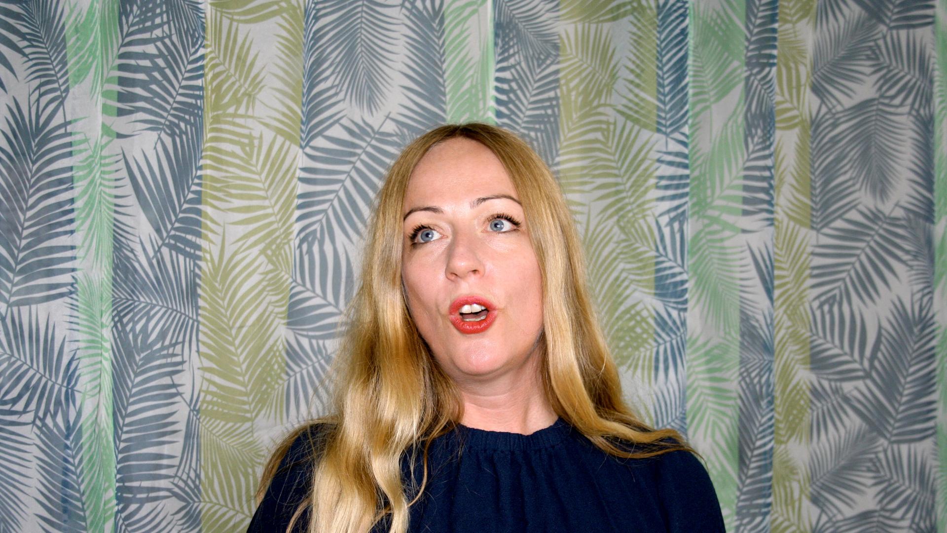 Die Profi-Jodlerin Doreen Kutzke formt den Mund zum Gesang. Sie hat lange blonde Haare und trägt eine dunkelblaue Bluse. Sie steht vor einem grünlich gemusterten Vorhang.