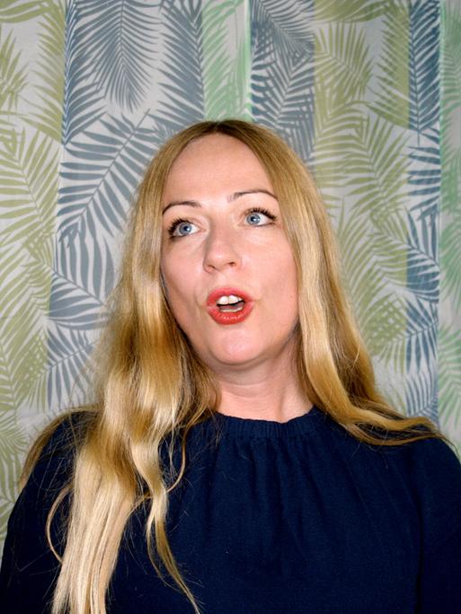 Die Profi-Jodlerin Doreen Kutzke formt den Mund zum Gesang. Sie hat lange blonde Haare und trägt eine dunkelblaue Bluse. Sie steht vor einem grünlich gemusterten Vorhang.