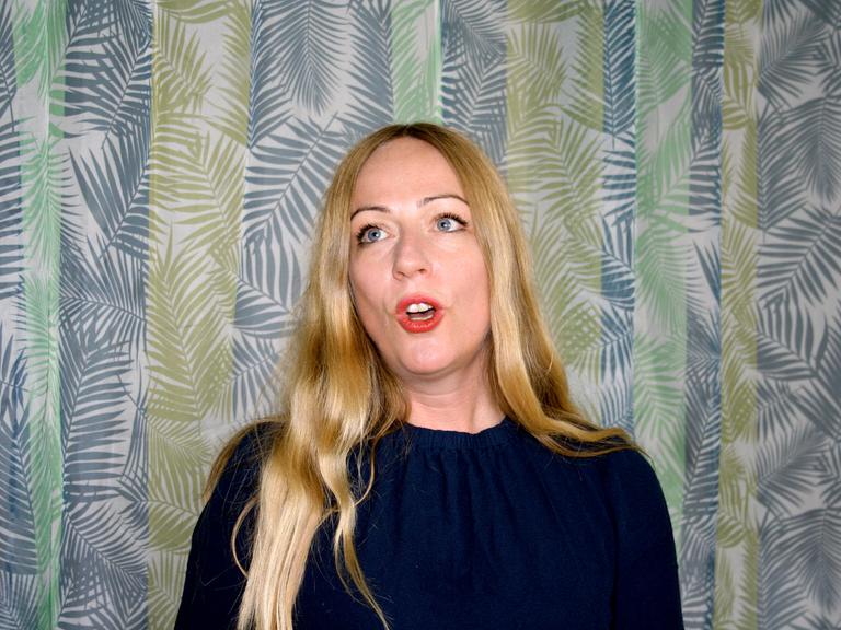 Die Profi-Jodlerin Doreen Kutzke formt den Mund zum Gesang. Sie hat lange blonde Haare und trägt eine dunkelblaue Bluse. Sie steht vor einem grünlich gemusterten Vorhang.