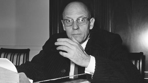 Foto (schwarz-weiß) des französischen Philosphen Michael Foucault, der an einem Schreibtisch sitzt, in die Kamera schaut und gestikuliert,
