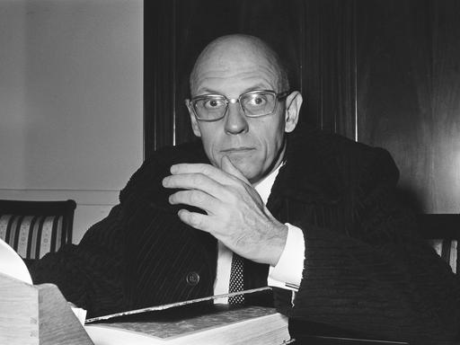 Foto (schwarz-weiß) des französischen Philosphen Michael Foucault, der an einem Schreibtisch sitzt, in die Kamera schaut und gestikuliert,