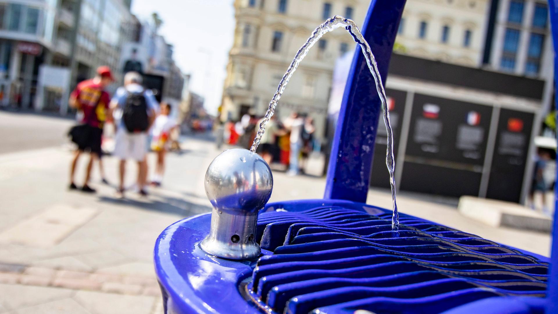 Trinkbrunnen bei Checkpoint Charlie in Berlin, im Hintergrund sind zwei Menschen mit Umhängetasche und Rucksack zu sehen