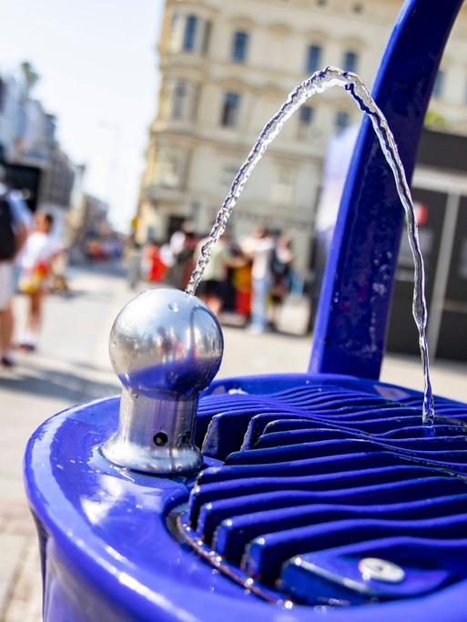 Trinkbrunnen bei Checkpoint Charlie in Berlin, im Hintergrund sind zwei Menschen mit Umhängetasche und Rucksack zu sehen