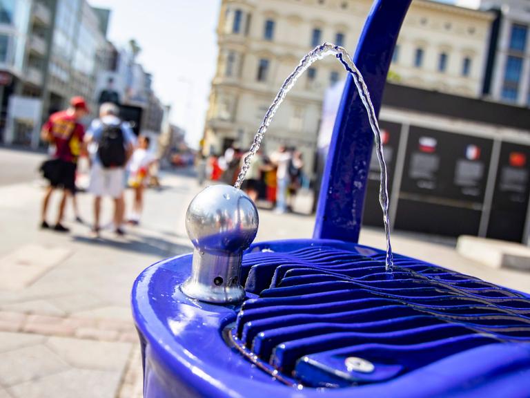 Trinkbrunnen bei Checkpoint Charlie in Berlin, im Hintergrund sind zwei Menschen mit Umhängetasche und Rucksack zu sehen