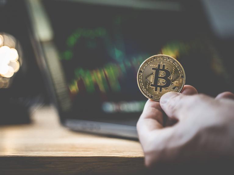 Eine physische goldene Bitcoin vor einem Computermonitor