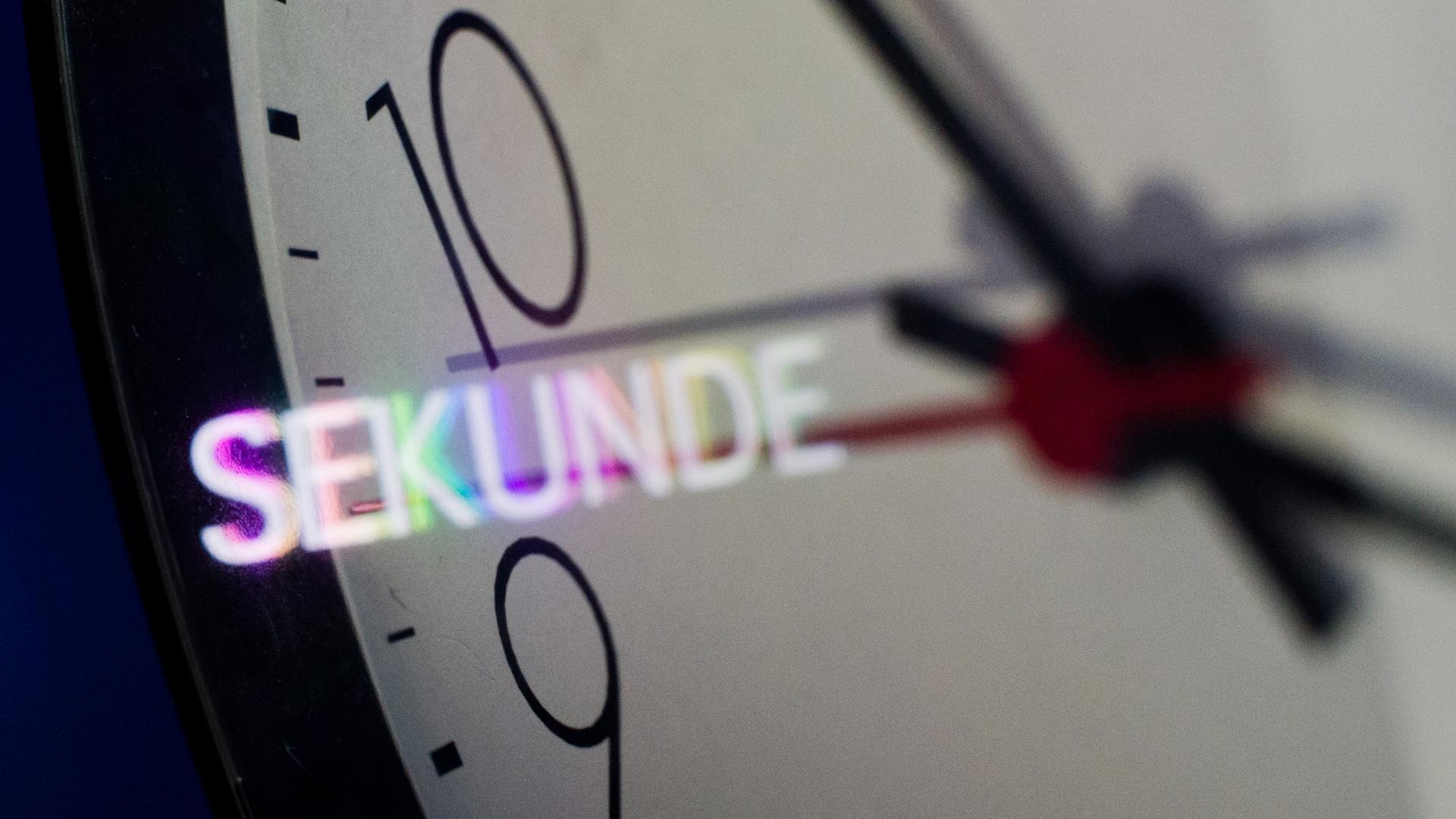 Nahaufnahme eines Ziffernblatts einer Wanduhr. Das Wort "Sekunde" spiegelt sich auf dem Ziffernblatt.