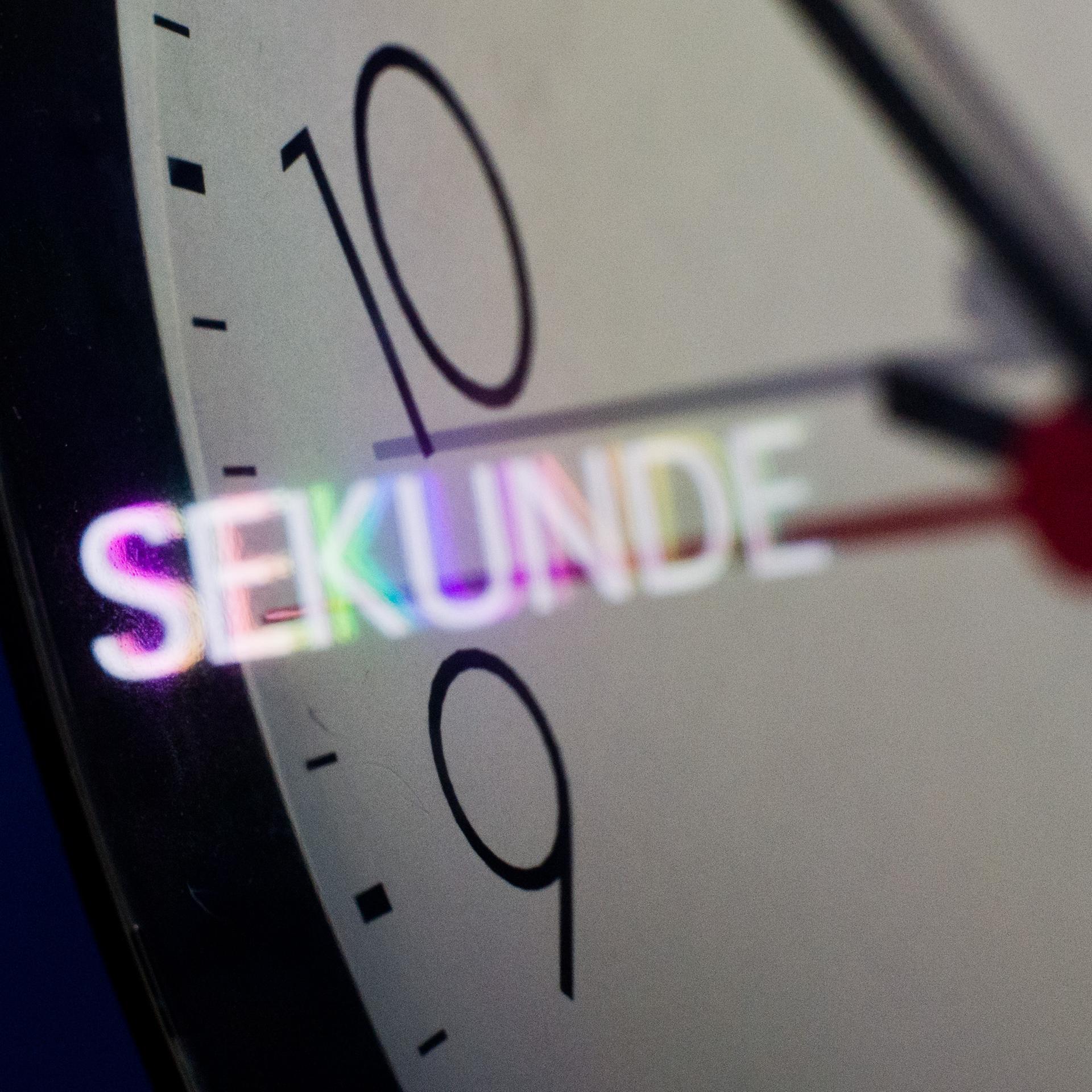 Nahaufnahme eines Ziffernblatts einer Wanduhr. Das Wort "Sekunde" spiegelt sich auf dem Ziffernblatt.