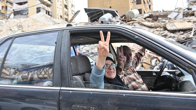 Eine Frau und ein Mann sitzen in einem Auto. Beide machen eine Geste mit der Hand.